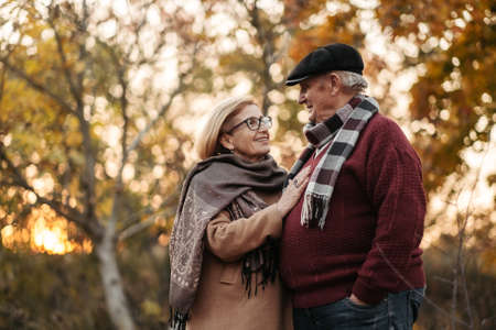Elderly man lovingly looks at his elegant pensioner, hugs and enjoys her beauty, senior married couple dancing in autumn park, celebrating anniversary, affectionの写真素材
