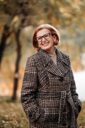Beautiful and slim elderly woman in a woolen coat, scarf posing, standing under a beautiful tree with golden leaves in an autumn park, relaxing and enjoying the bright foliage, happy retirementの写真素材