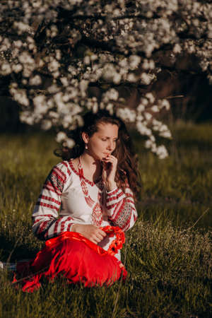 Walks in nature in the countryside. Close-up portrait of a young woman in a blooming garden. A girl in a national costume, a bandage with a flower on her head, an embroidered shirt and a red skirt.の写真素材