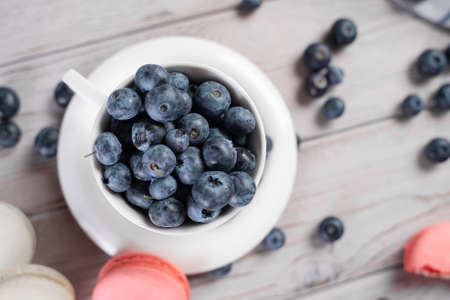 Top view of ceramic cup of ripe blueberries placed on table near sweet macaroons in morning