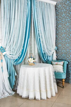 Comfortable corner of room in classic design with blue and white curtains, bouquet of flowers on the table with white tablecloth, an armchair with pillow at window.の写真素材