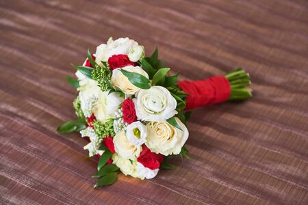Beautiful wedding bouquet close-up with white, red roses, greens and other flowers with red ribbon on brown fabric background.の写真素材