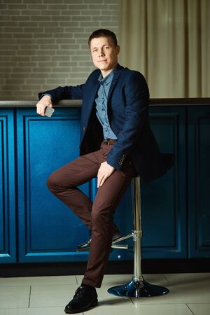 A young man sits on a bar stool in full growth, leaning on a counter, mobile in hand.の写真素材