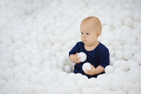 Cute little baby boy sits plays in dry pool of white balls, looks away.の写真素材