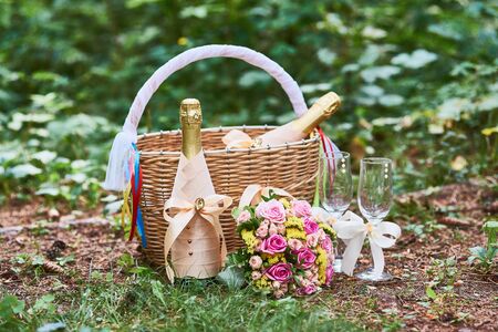 Beautiful composition of wicker basket with colored satin ribbons, two decorated champagne bottles, romantic wedding bouquet, wine glasses on the grass in forest on green background.の写真素材