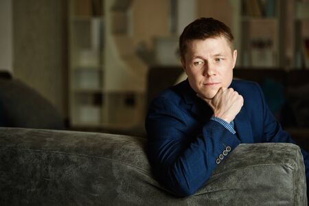 Young man leaning against back of the sofa, looking down thoughtfully, propping his chin with his hand.の写真素材