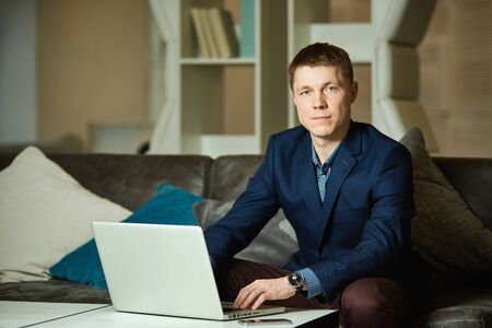 Portrait of a young handsome businessman, a man sitting at a table and typing on a laptop, looking at the camera.の写真素材