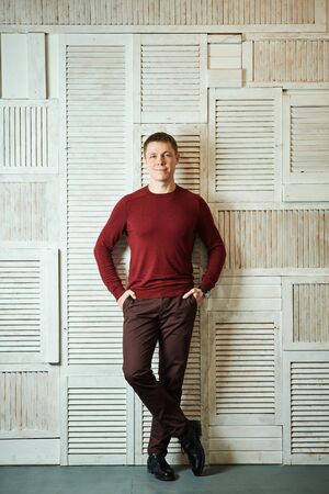 Handsome young man is standing full-length with his legs crossed leaning against wall with his back holding his hands in his pockets, smiling, looking at camera.の写真素材