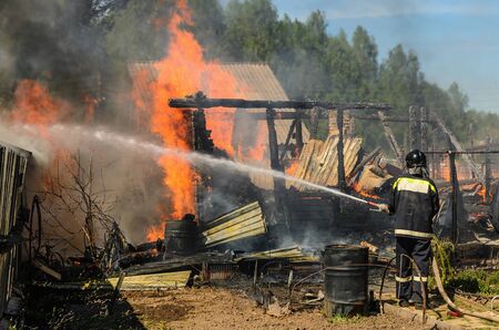 Firefighter in protective suit and helmet extinguishes a burning house engulfed in flames using fire hose with water. Fire in action. Fire fighting.の写真素材