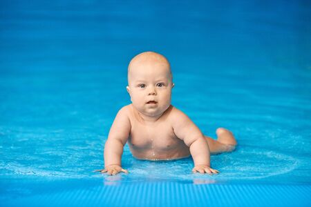 Little baby boy lying in blue swimming pool in water. Concept active lifestyle of child. Kid looks at the camera.の写真素材