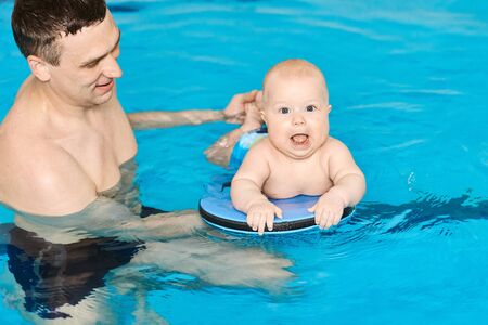 Happy little baby boy swimming in water pool with help from hands father and board. Swim lessons for children. Healthy Family Life. Active family spending leisure.の写真素材