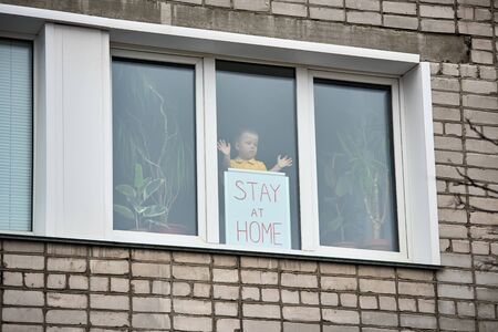 Child boy stands on window behind Board with inscription Stay at home, leaning on glass, looks to left. View from street. Stay at home for quarantine prevention of coronavirus pandemic.の写真素材