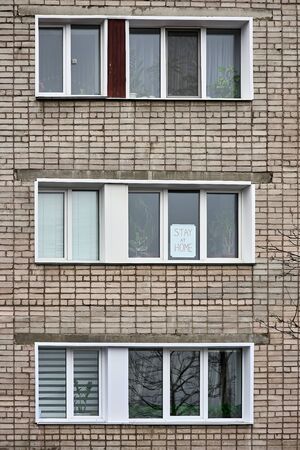 Banner with inscription stay at home, stands behind the glass on window of brick house. View from the street. Call for quarantine prevention of coronavirus pandemic.の写真素材