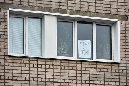 Banner with inscription stay at home, stands behind the glass on window of brick house. View from the street. Call for quarantine prevention of coronavirus pandemic.の写真素材