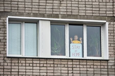 Child boy stands on windows behind banner with inscription Stay at home, leaning on glass, looks to right. View from street. Stay at home for quarantine prevention of coronavirus pandemic.の写真素材