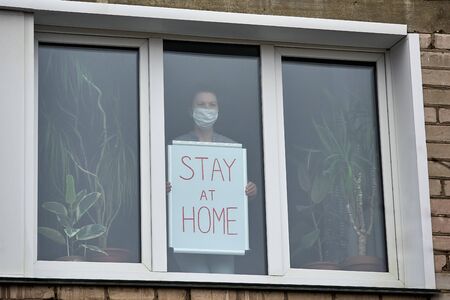 Woman in medical mask is at home, standing outside window, holding board with inscription stay at home. Call for self-isolation. View from street. Close-up.の写真素材