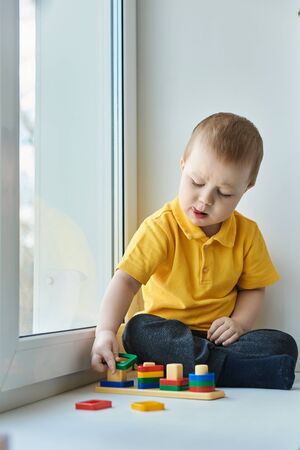 Small child boy is sitting at home on the windowsill, playing a developmental game and looking out the window. Home development of a child in isolation.の写真素材