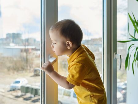 Adorable little child in yellow shirt is standing on window at home, holding the handle and looking out the window at the street.の写真素材