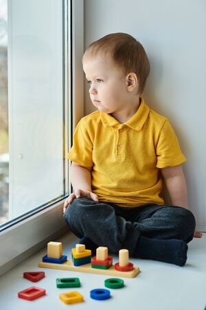Small child boy is sitting at home on the windowsill, playing a developmental game and looking out the window. Concept of early development of children.の写真素材