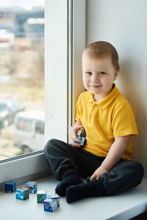 Cute little boy is sitting at home on the windowsill, playing cubes, smiling and looking at the camera.の写真素材