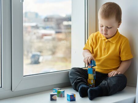 Beautiful little child boy is sitting at home on the window, enthusiastic playing with cubes and building a tower. Children home games in isolation.の写真素材
