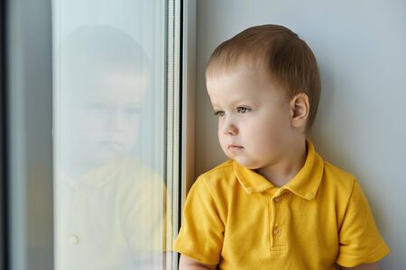 Cute little child in a yellow shirt sits alone at home on the windowsill and looks out window at the street.の写真素材