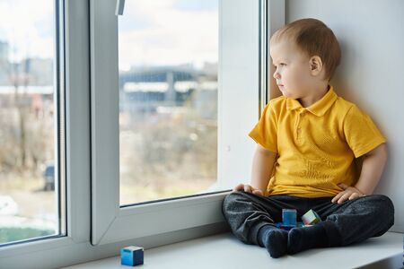 Small child in yellow shirt sits at home on windowsill, playing cubes and watching out the window.の写真素材