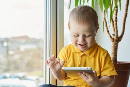 Cheerful little boy sitting on windowsill next to window, holding tablet computer and tapping the screen with his finger. Children entertainment at home.の写真素材