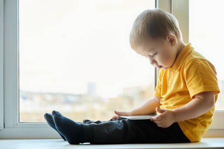 Child boy in yellow shirt sits on windowsill at the window and watches video cartoons on the screen of tablet computer.の写真素材