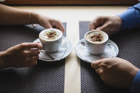 Man and woman sit at table in cafe and hold two cups of coffee in their hands.の写真素材