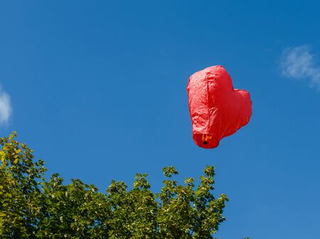 Chinese lighted red paper lantern in shape of heart soars high into blue sky against the green foliage of trees.の写真素材