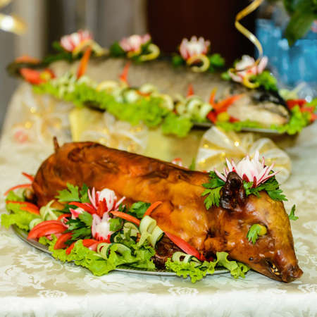 Young roast pig is lying beautifully on a dish decorated with fresh vegetables and herbs.の写真素材