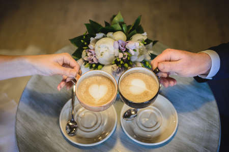 Two cups of coffee in the hands of man and woman, raised above the table against background of beautiful bouquet of flowers.の写真素材