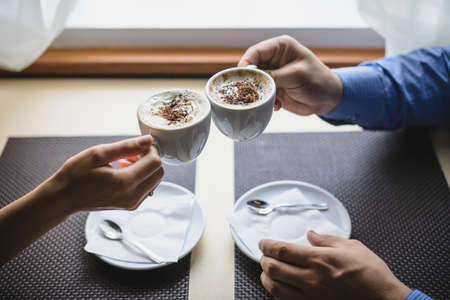 Two cups of coffee in the hands of man and woman are raised above the table and touch each other.の写真素材