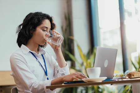 Young woman drinking water while typing on laptop during lunch break in bright office cafeteriaの写真素材