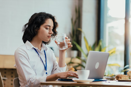 Young woman drinking water while typing on laptop during lunch break in bright office cafeteriaの写真素材