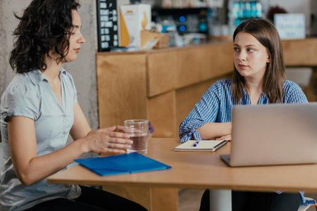 Two female coworkers discussing project over coffee in office cafeteria