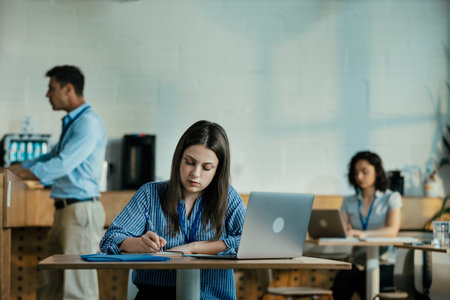 Female Student Taking Notes from Online Lecture in Sunny University Cafeteria During Lunchの写真素材