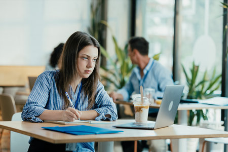 Female Student Taking Notes from Online Lecture in Sunny University Cafeteria During Lunchの写真素材