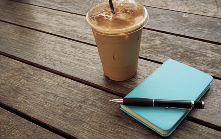 Ice coffee in takeaway cup with notebook and pen on the side. Book and pen with ice latte on wood...