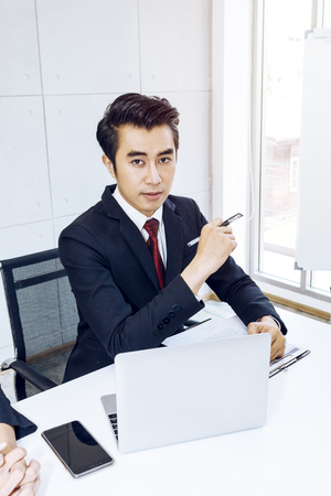 Young handsome businessman smiling and smart with pen and laptop at office.He is 20-30 years old in black suit.Copy space.の写真素材
