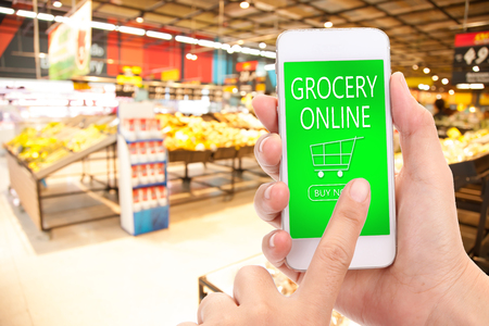 Woman hand click BUY NOW on mobile with blur supermarket background, Grocery online,  delivery...