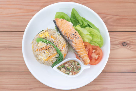 Salmon steak, egg fried rice, fish sauce with chili, Butterhead Lettuce.  and tomatoes in a ceramic plate on a pine wood table.の写真素材