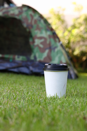 hot coffee drink, white disposable cup on green grass lawn campsite, tent camping trip journey travel in natureの写真素材