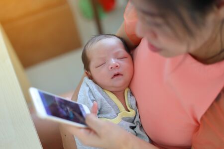 mother playing mobile phone in business home office with cute baby newborn sleeping