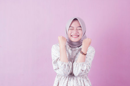 Smiling happy young woman in hijab wearing floral shirt doing winner gesture celebrating fist saying...