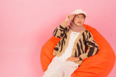 beautiful young woman in glasses and hat sitting on orange bean bag with angle from above, pink isolated studio backgroundの写真素材