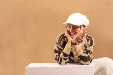 beautiful young woman in glasses and hat sitting with hand supporting her chin smiling sweetly, brown backgroundの写真素材