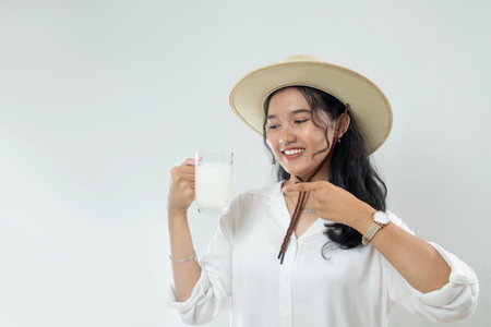 Beautiful young Asian woman in hat and holding glass of milk with other hand pointing, healthy lifestyle with drinking milk, cow's milk, soy milk for calcium, isolated white backgrの写真素材