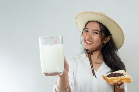 Beautiful young Asian woman with straw hat showing milk in her glass with one hand holding bread, woman having breakfast concept, isolated white backgroundの写真素材